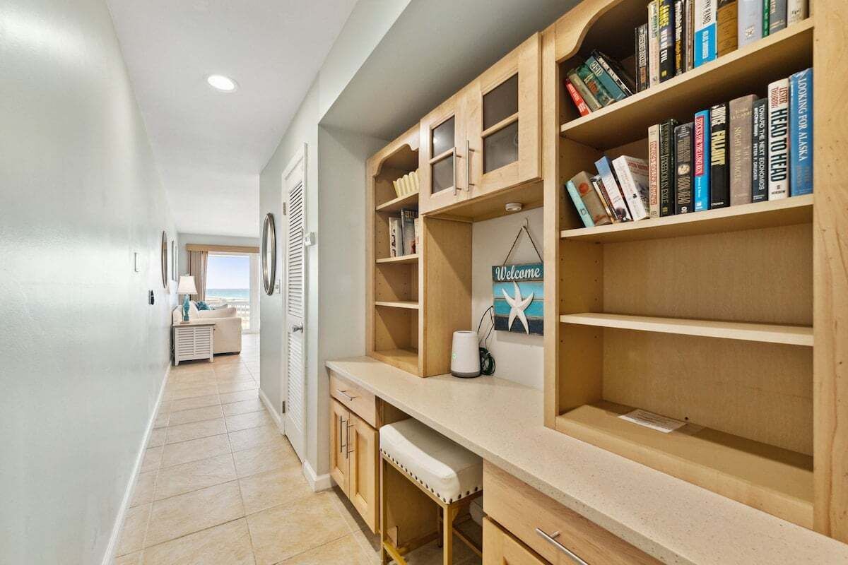 A hallway with a wooden desk and bookshelves, a white wall, and a window with a view.
