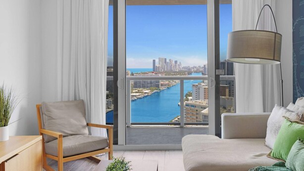 A modern living room with a large window, a sofa, a chair, and a floor lamp. - Infinite View| Balcony| Pool+ Gym (Hallandale Beach)