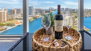 A bottle of wine and two glasses on a woven tray with a view of a cityscape and water. - Infinite View| Balcony| Pool+ Gym (Hallandale Beach)