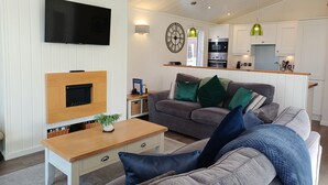 Una sala de estar moderna con televisor de pantalla plana, una mesa de centro de madera y un sofá gris con cojines verde y azul. Se ve una cocina con gabinetes blancos y horno empotrado al fondo.