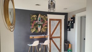 Una habitación con una pared oscura, una puerta blanca, un escritorio de madera, dos sillas blancas, un adorno en la pared y una lámpara en el techo.
