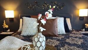 Una habitación de hotel con una cama, almohadas decorativas, un jarrón con flores y apliques de pared.