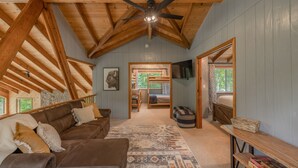 A cozy cabin interior with a high ceiling, wooden beams, a ceiling fan, a large window, a sofa, a coffee table, and a TV. - Alpine Woods by Innsbrook Vacations! (Innsbrook)