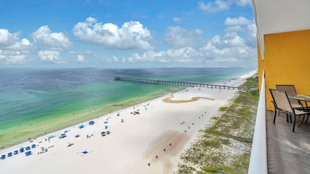 A beach view with a pier extending into the ocean, a sandy beach with umbrellas, and a balcony with chairs.