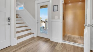 A clean, modern hallway with a wooden staircase, a glass door leading to an outdoor area, and a wooden elevator. - Vitamin Sea: Coastal Chic Home with Private Pool, Elevator (Oak Island)