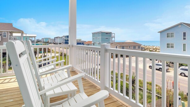 A wooden deck with white chairs, a white railing, and a view of a residential area with houses and a road. - Vitamin Sea: Coastal Chic Home with Private Pool, Elevator (Oak Island)