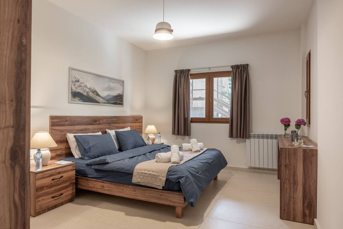 A bedroom with a wooden bed, nightstands, a window with curtains, a radiator, and a wall-mounted shelf with flowers.