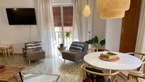 A modern living room with a flat-screen TV, two sofas with striped pillows, a round dining table with a woven placemat, and a large plant.
