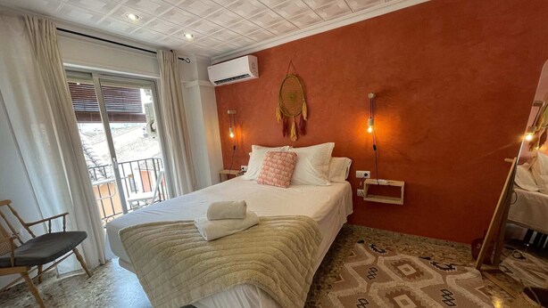 A hotel room with a bed, a chair, an air conditioning unit, a wall-mounted light, and a patterned rug.