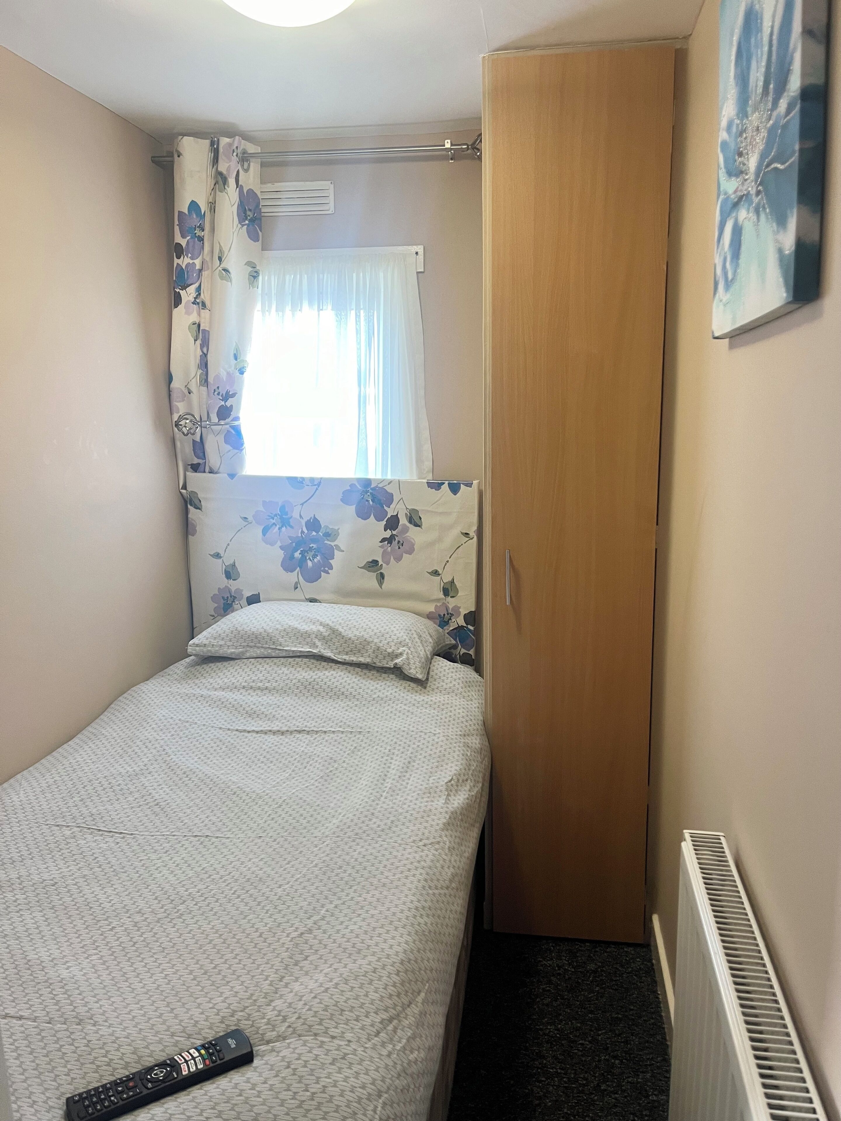 A small bedroom with a single bed, a wooden wardrobe, a radiator, and a remote control on the bed.