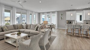Ein modernes Wohnzimmer mit einem Ecksofa, einem Couchtisch, Korbstühlen, einem Essbereich mit Tisch und Stühlen sowie einer Küche mit weißen Schränken und Barhocker.