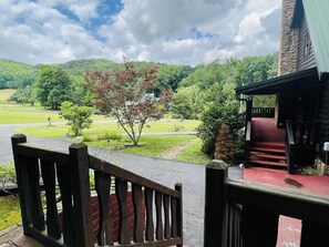 A wooden deck with a stone wall, a stone building with a red roof, and a view of a green landscape with trees and a hill. - Burning Tree 3 Bedroom Cabin by RedAwning (Sevierville)