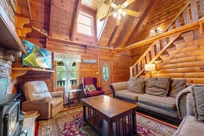 A cozy log cabin interior with a fireplace, wooden furniture, a ceiling fan, and a staircase. - Burning Tree 3 Bedroom Cabin by RedAwning (Sevierville)