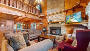 A cozy cabin interior with a fireplace, wooden walls, a sofa, a red armchair, and a ceiling fan.