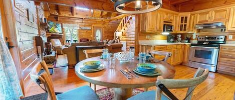 A wooden cabin interior with a dining area, kitchen, and a staircase leading to a loft bedroom.