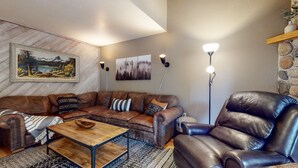 Un salón con un sofá de cuero marrón, una mesa de centro de madera y un sillón de cuero negro. Hay dos lámparas de pie fijadas a la pared y un cuadro de paisaje enmarcado sobre el sofá.