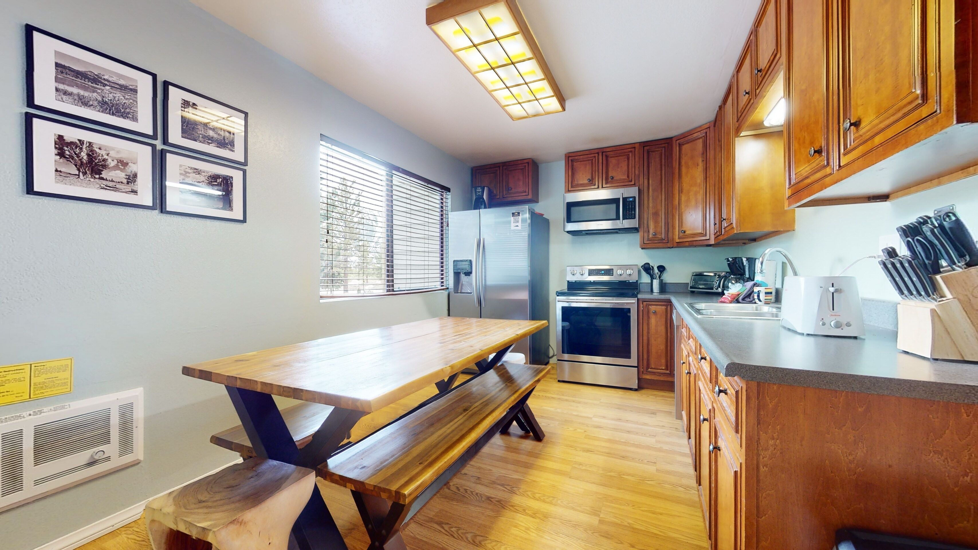 A kitchen with wooden cabinets, a stainless steel refrigerator, a microwave, an oven, and a sink. There is a wooden table and bench, and framed pictures on the wall.