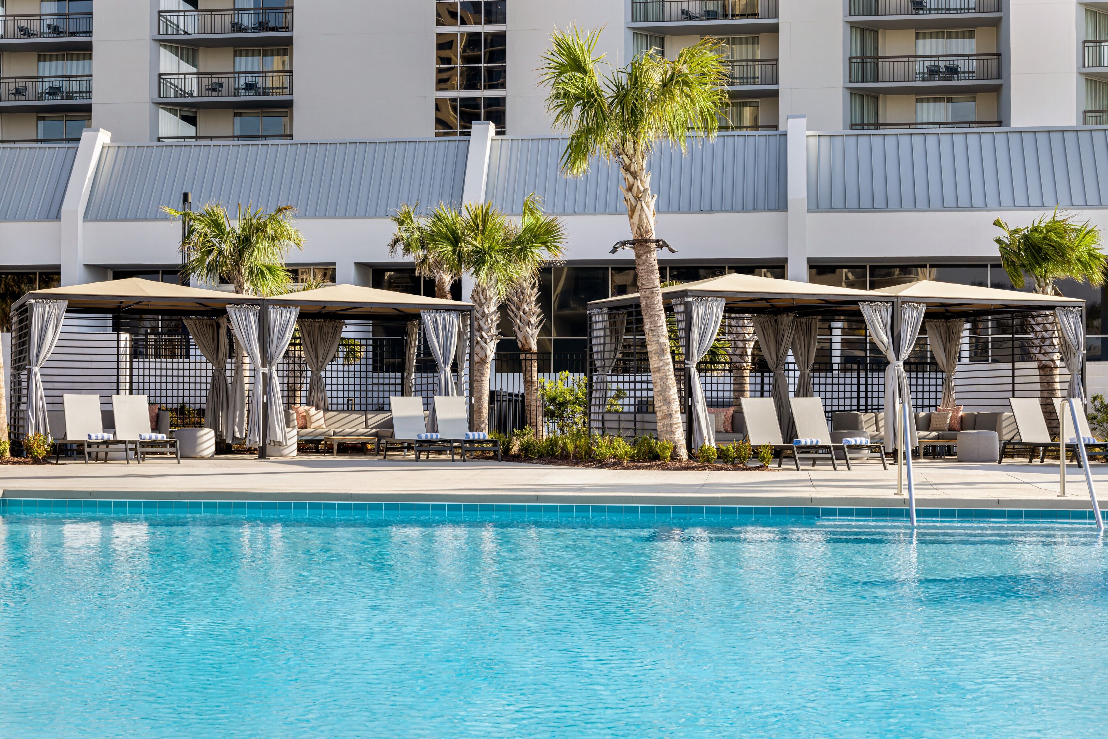 A swimming pool with lounge chairs and umbrellas, surrounded by palm trees and a modern building with balconies.