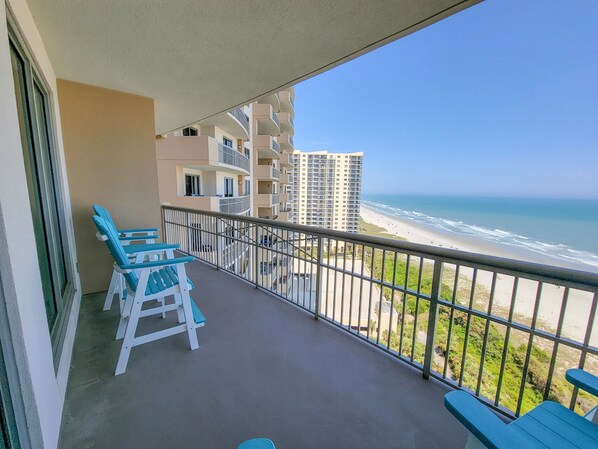 A balcony with blue chairs and a table, overlooking a beach and high-rise buildings. - 1104 Margate 3 Bedroom Condo (Myrtle Beach)