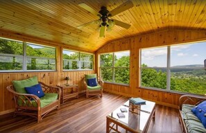 A wooden-paneled room with wicker furniture, a glass coffee table, and large windows offering a view of greenery. - Little Greenbriar Lodge 13 Bedroom Cabin by RedAwning (Sevierville)
