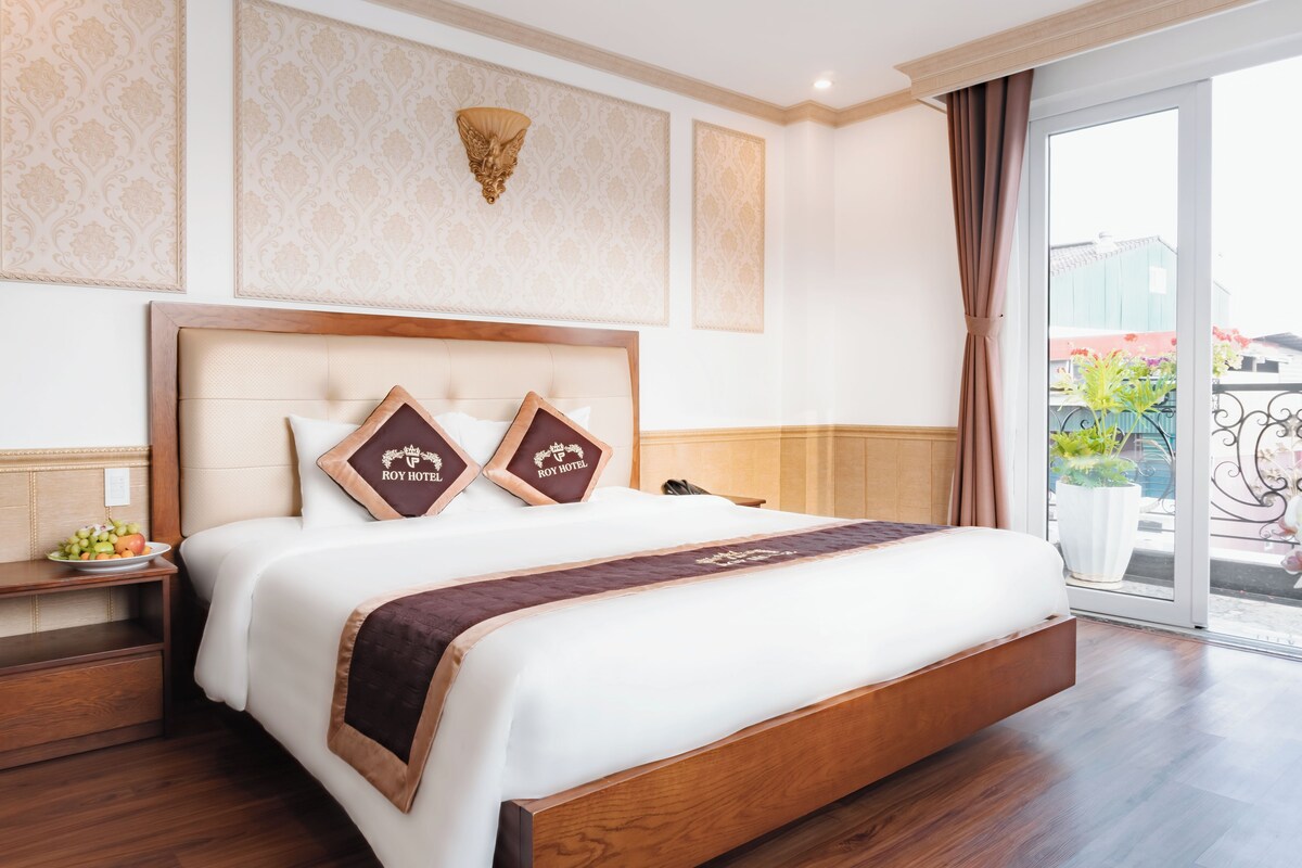 Habitación de hotel con una cama grande, una mesita de noche con frutero, una lámpara de pared y vistas al exterior.