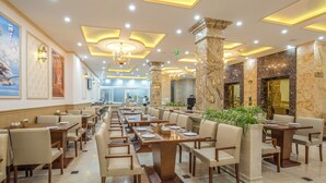 Un restaurante espacioso con largas mesas y sillas, una columna de mármol y una araña.