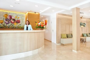 A reception area with a wooden reception desk, a painting of flowers, a seating area with cushions, and a wall-mounted light fixture. - Universal Hotel Laguna (Capdepera)