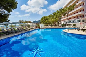A swimming pool with lounge chairs and umbrellas, surrounded by a building with balconies and trees.