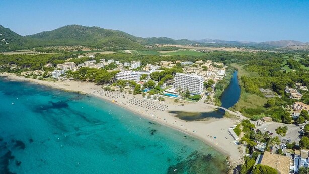 An aerial view of a coastal resort with a beach, hotel buildings, and a river. - Universal Hotel Laguna (Capdepera)