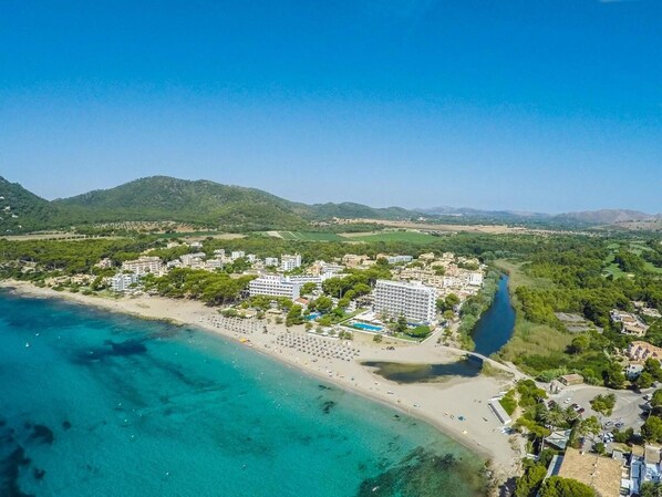Vista aérea de um resort à beira-mar, com praia, edifícios de hotel e um rio.