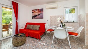 A compact room with a red sofa, a dining table with chairs, a wicker side table, and a view of greenery outside. - Apartment OLIVE with private garden 250m2 (Vinkuran)