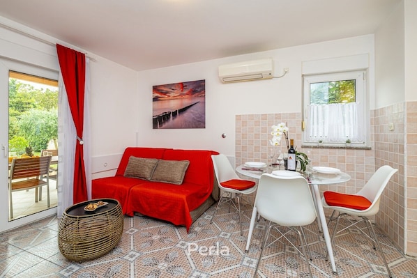 A compact room with a red sofa, a dining table with chairs, a wicker side table, and a view of greenery outside. - Apartment OLIVE with private garden 250m2 (Vinkuran)