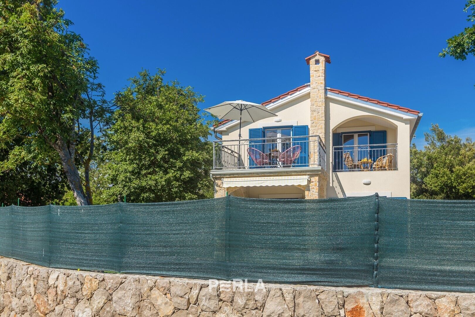 Une maison à deux étages avec un balcon, un mur de pierre au premier plan et un ciel bleu dégagé.