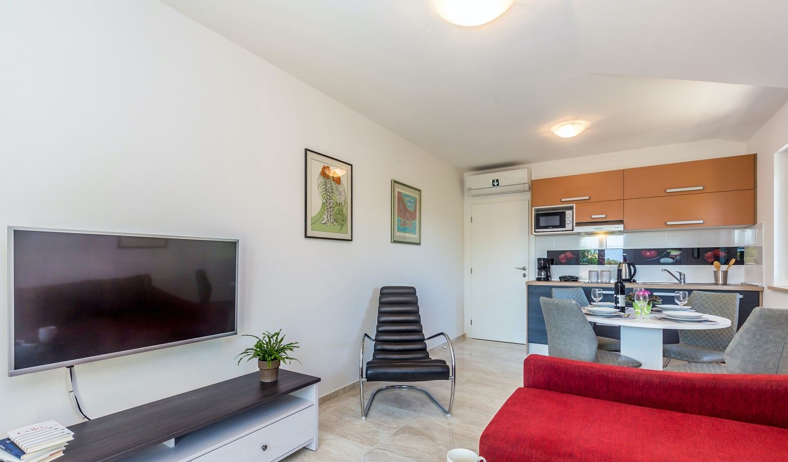 Ein modernes Wohnzimmer mit Flachbildfernseher, einem roten Sofa, einem Esstisch mit Stühlen und einer Küchenzeile mit Schränken und einer Mikrowelle.