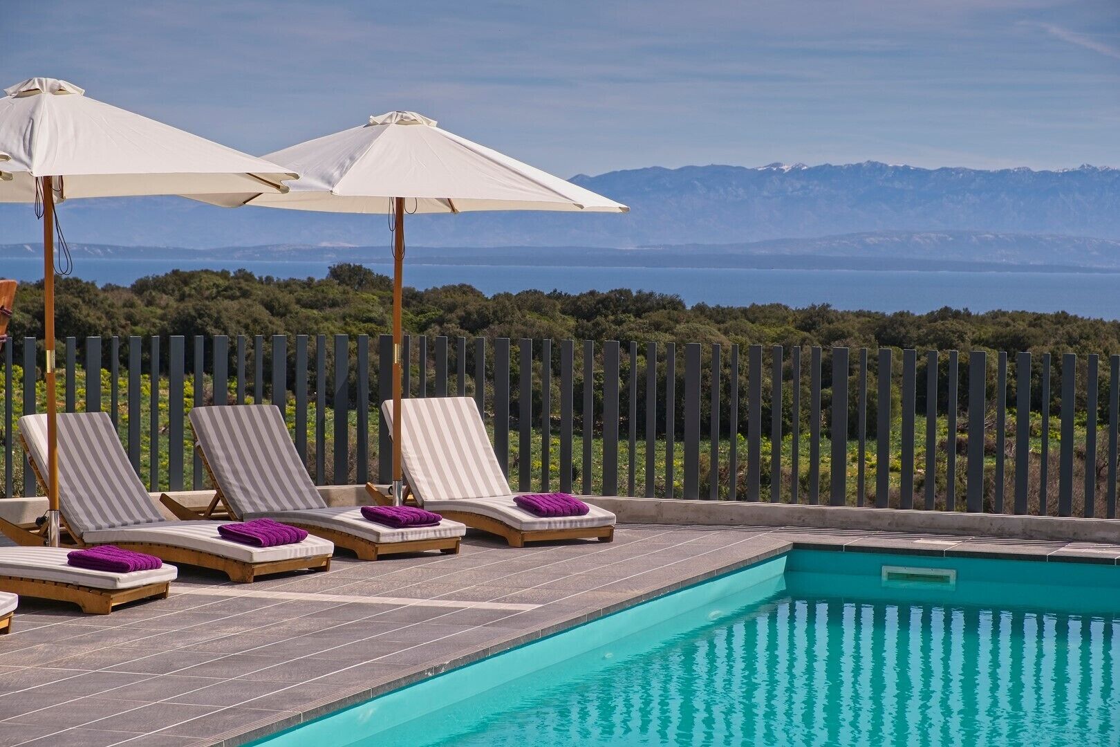 A poolside area with lounge chairs and umbrellas, a clear view of the sea and mountains in the background.