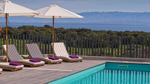 Un espace aménagé au bord de la piscine, avec des chaises longues et des parasols, offrant une vue dégagée sur la mer et les montagnes en arrière-plan.