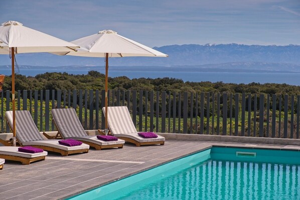 A poolside area with lounge chairs and umbrellas, a clear view of the sea and mountains in the background.
