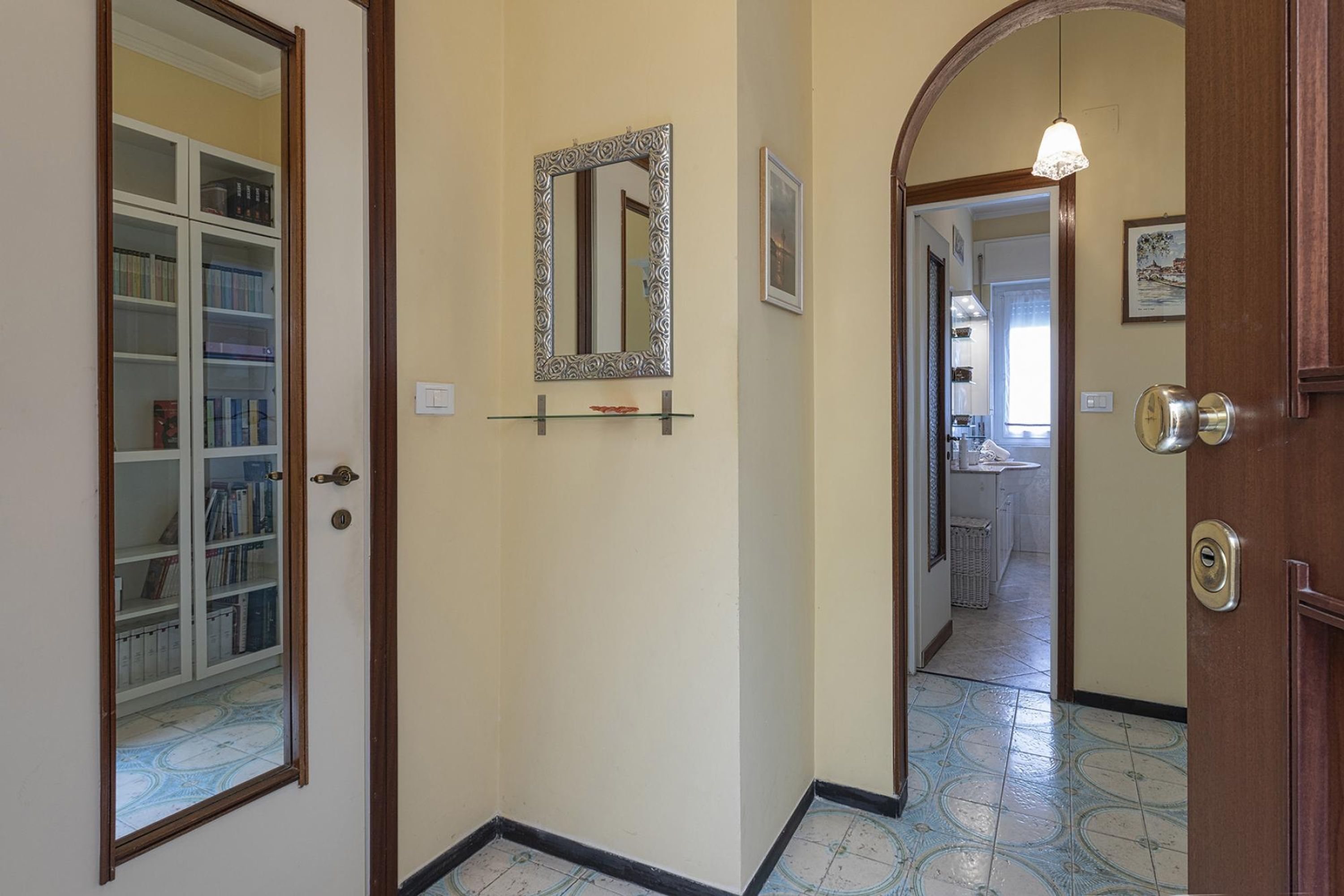 A hallway with a bookshelf, a decorative mirror, and a door leading to another room.