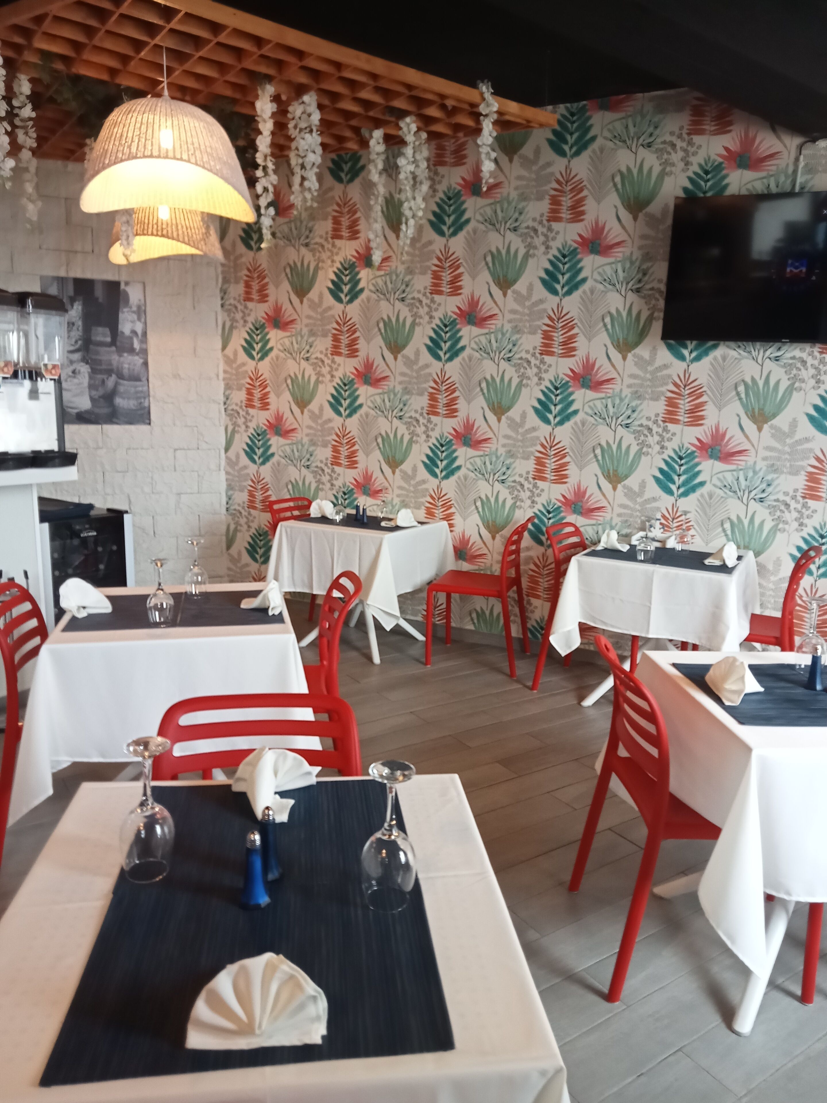 Interior de restaurante con mesas preparadas para comer, sillas rojas, una lámpara colgante y un papel tapiz estampado.