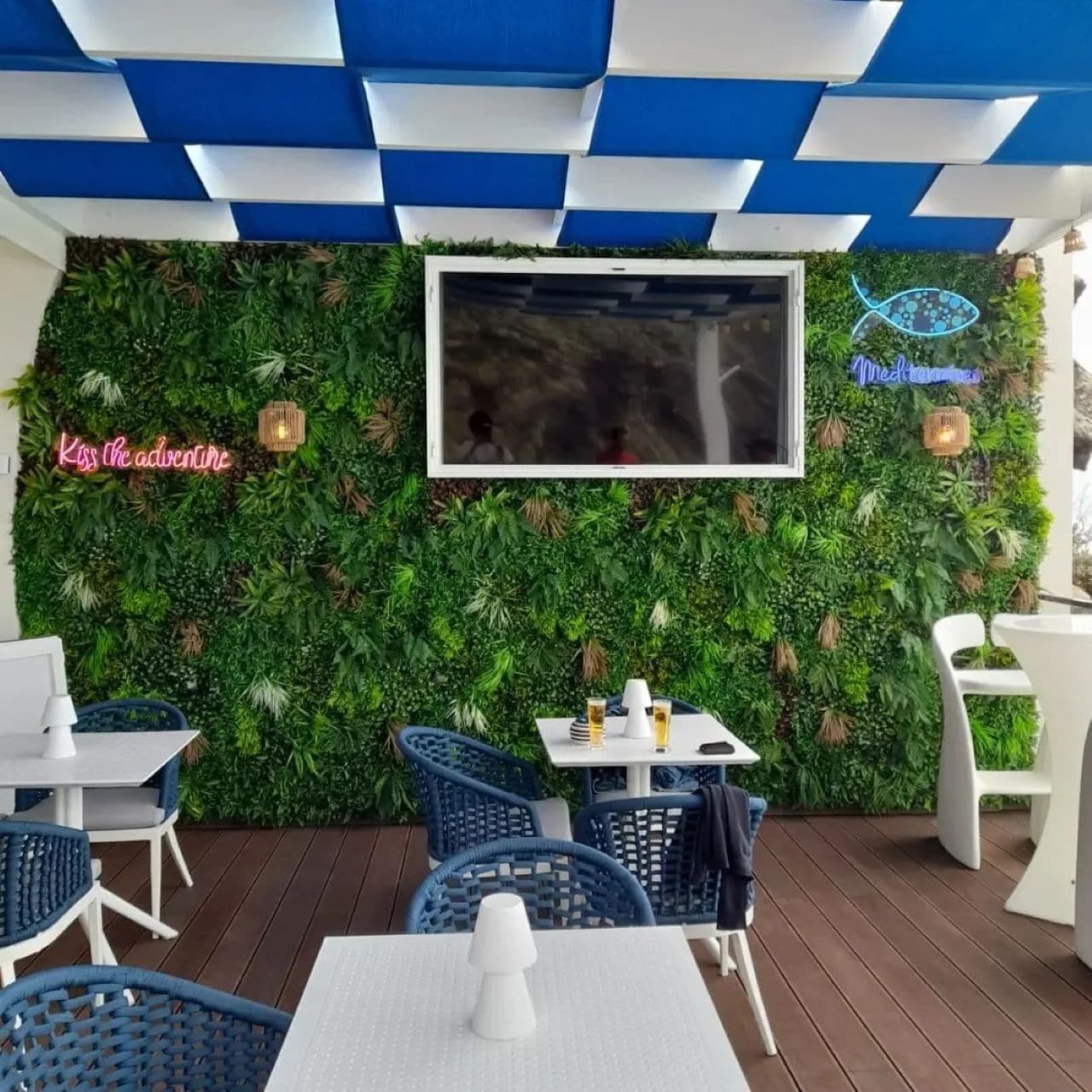 Un espace extérieur aménagé avec un mur végétal, un panneau lumineux au néon et un téléviseur fixé au mur.