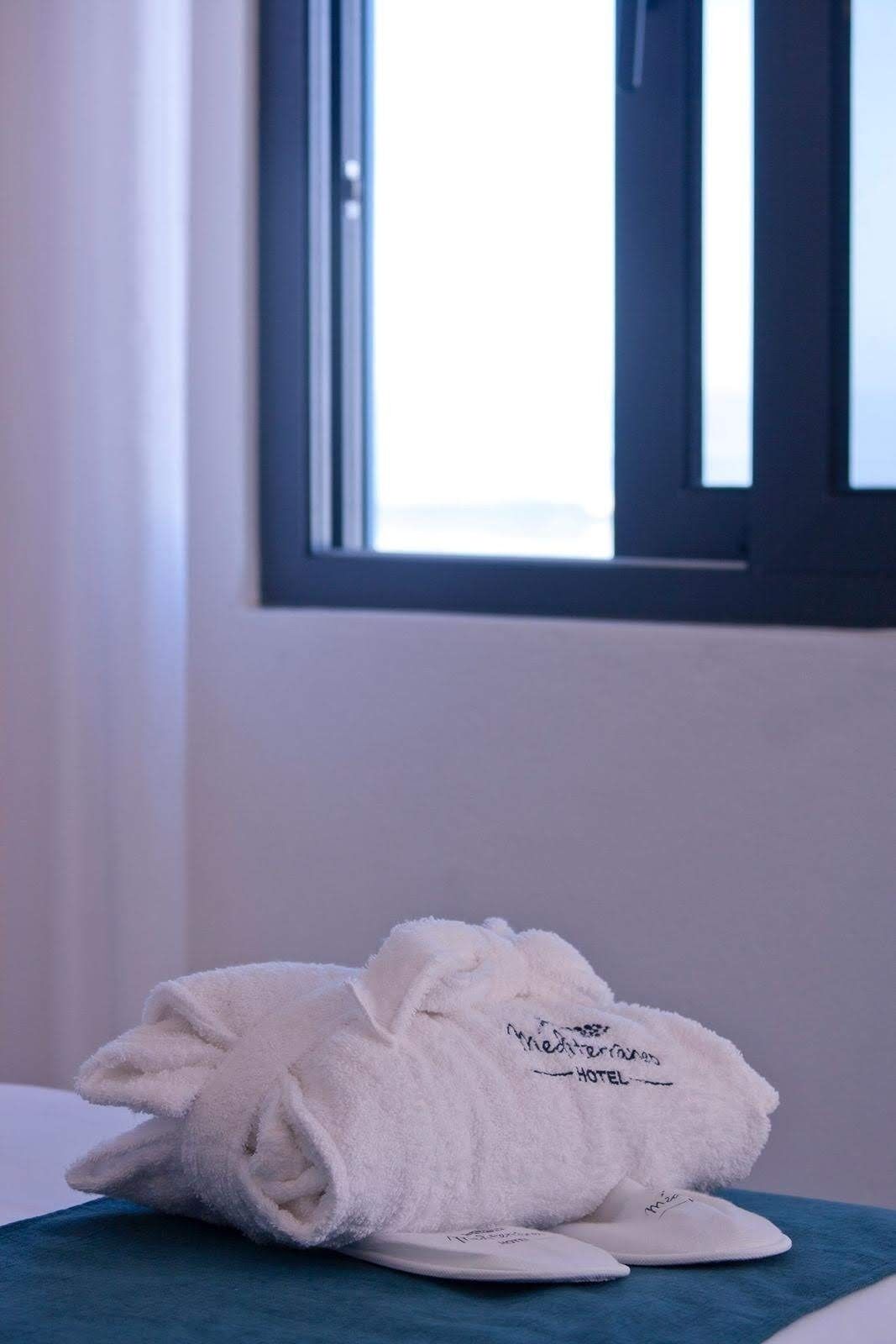A neatly folded white towel with "The Mediterranean Hotel" text on it, placed on a blue towel on a bed with white sheets.