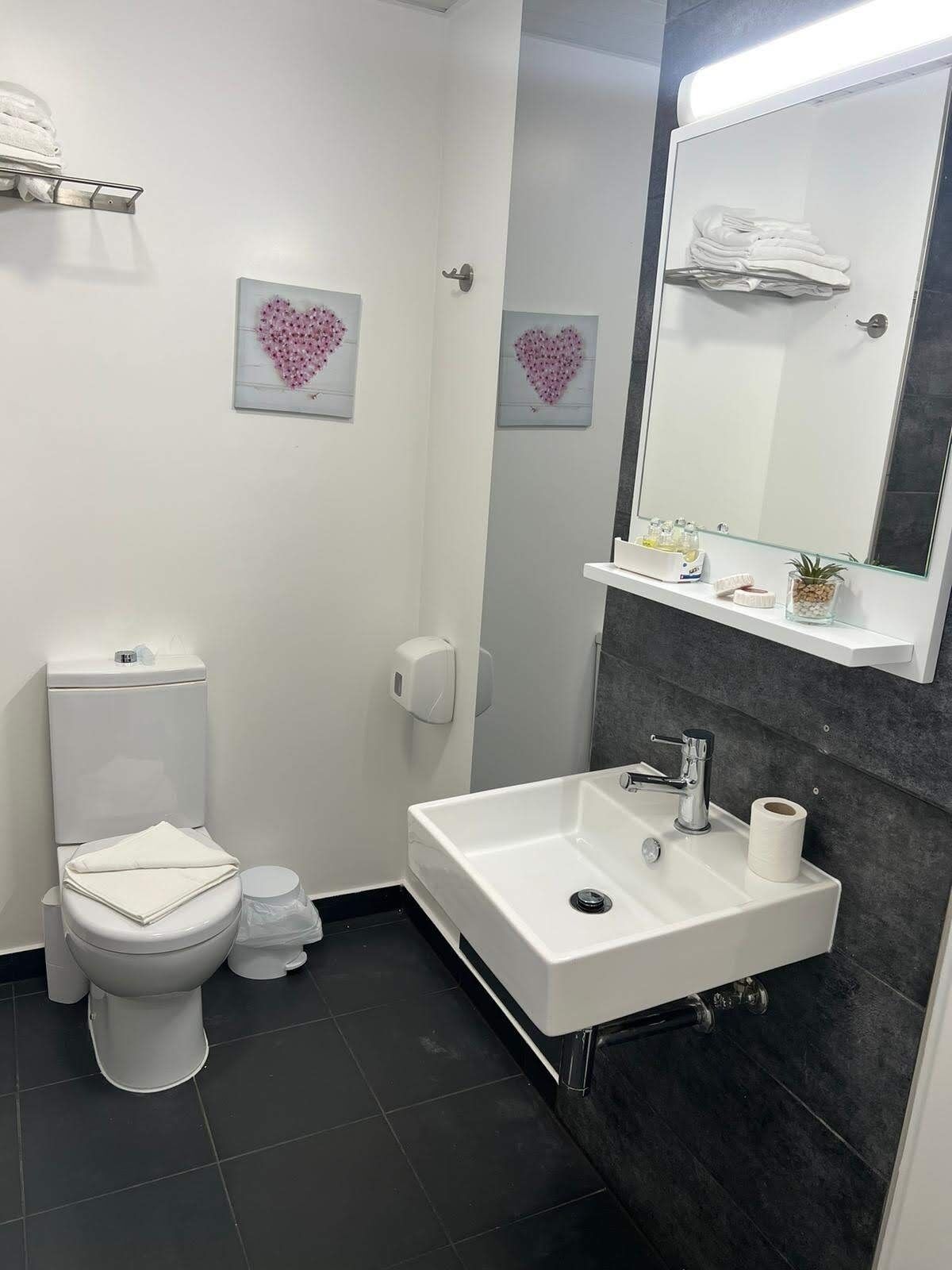 Ein modernes Badezimmer mit Toilette und Waschbecken in Weiß. Ein großes Spiegel und Wanddekorationen in Herzform. Eine kleine Ablagefläche mit Toilettenartikeln.
