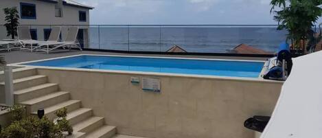 Una piscina con escalones de acceso, una zona cubierta con sillas de descanso y vistas al mar.