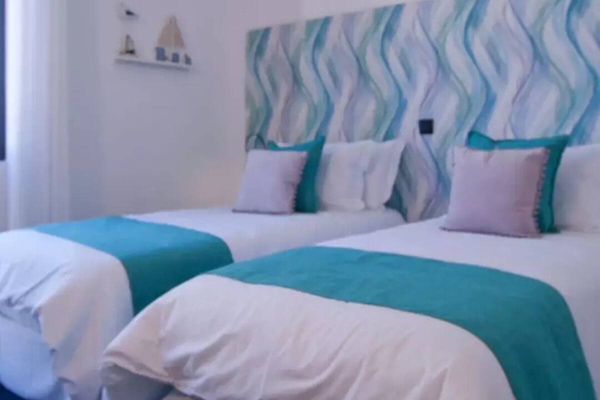 Hotellihuone, jossa on kaksi sänkyä. Kummassakin sängyssä on valkoiset vuodevaatteet ja turkoosit yksityiskohdat. Yhdellä seinällä on aaltoileva kuvio, ja huoneessa on pieni hylly, jossa on koriste-esineitä.