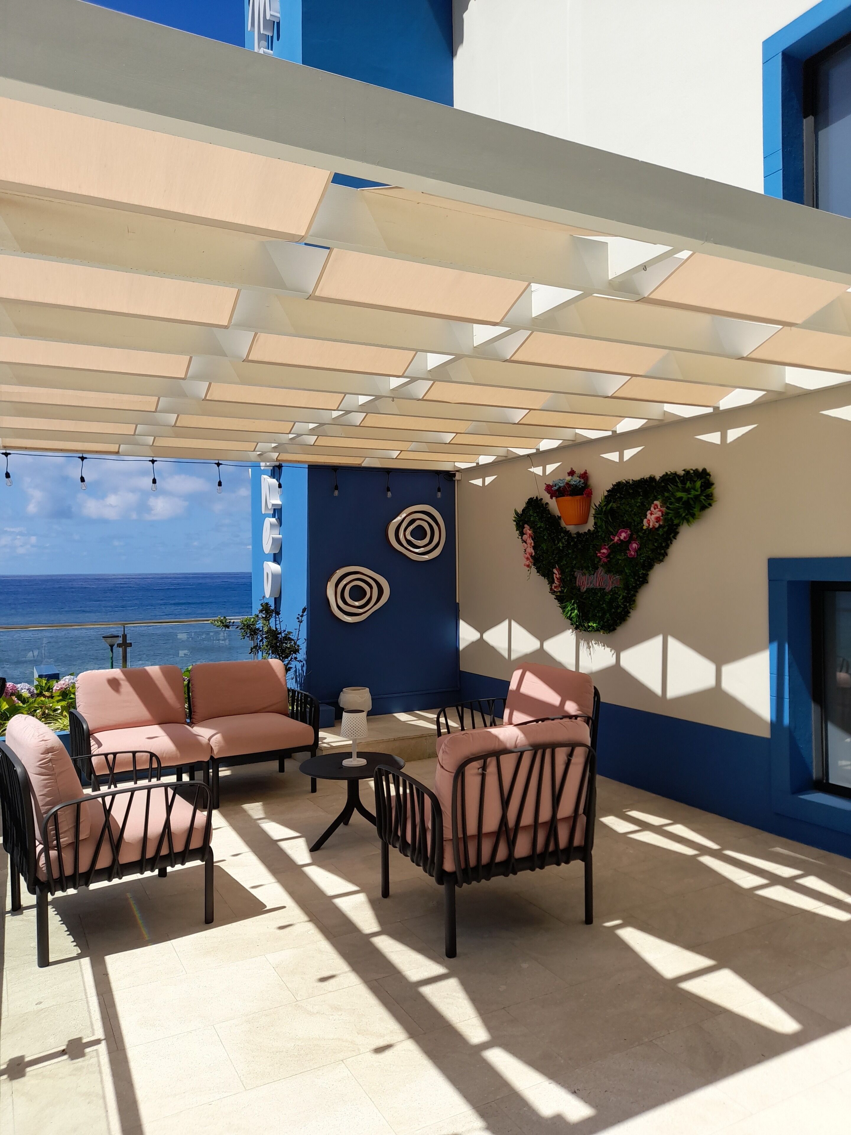 An outdoor seating area with a view of the sea, featuring a pergola, a wall with decorative circles, and a heart-shaped floral arrangement.
