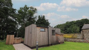 A wooden cabin with a small window and a door, surrounded by a grassy area and trees. - Brow Wood Cabin (Penrith)