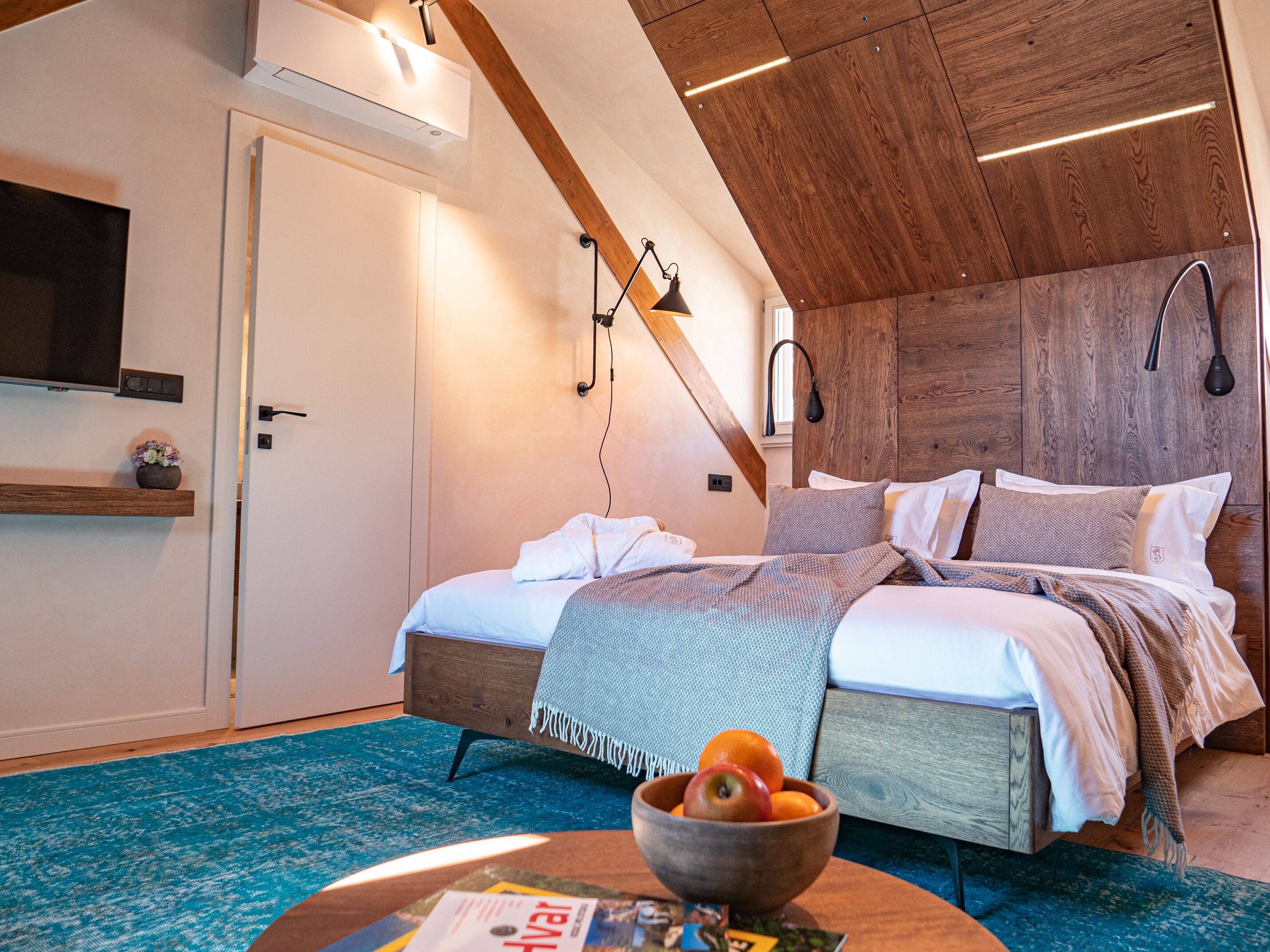 Ein modernes Schlafzimmer mit schrägem Dach, einem Bett mit vielen Kissen, einem hölzernen Nachttisch, einer Obstschale und einer Zeitschrift auf dem Boden.