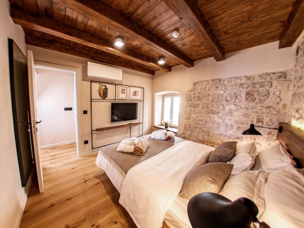 A bedroom with a stone wall, wooden ceiling beams, a bed with pillows, a TV mounted on the wall, and a window with a view. - Villa Porta Maestra (Hvar)
