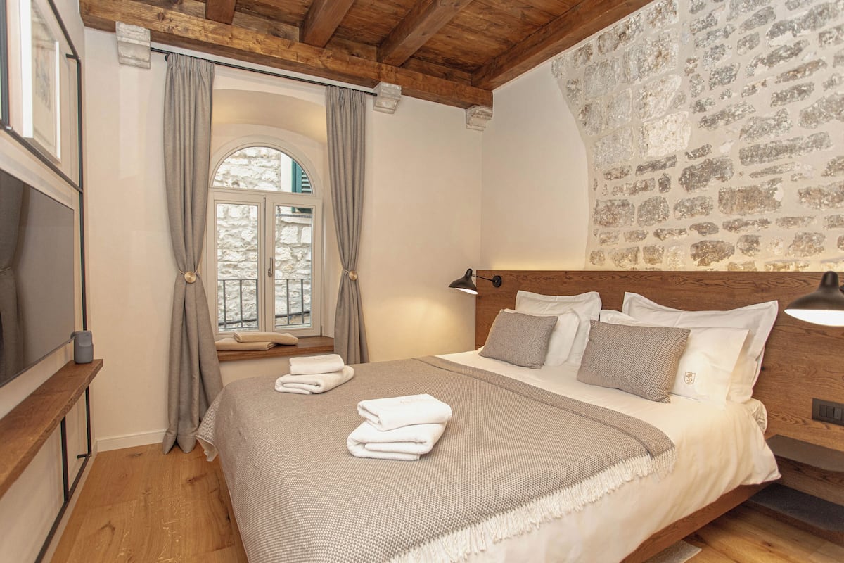 A bedroom with a wooden headboard, a stone wall, a large window with curtains, a bed with white linens, and a bedside table with a lamp.