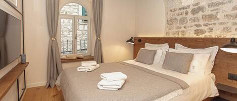 A bedroom with a wooden headboard, a stone wall, a large window with curtains, a bed with white linens, and a bedside table with a lamp.